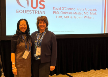 Dr. Christina Master and Dr. Kristy Arbogast stand next to each other in front of a black curtain, with a screen that has the US Equestrian logo projected, as well as the words "Safety Session."